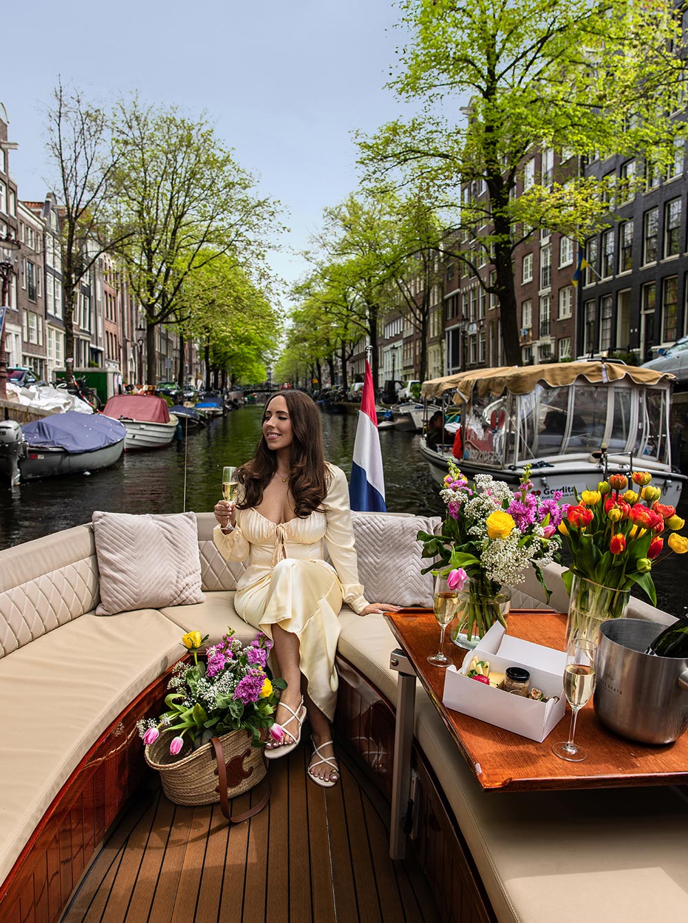 A woman in a cream dress sits on a luxury boat cruising an Amsterdam canal, holding a glass of champagne and surrounded by fresh tulip bouquets and a gourmet snack box. The scene features charming canal houses, docked boats, and spring greenery under a soft sky.