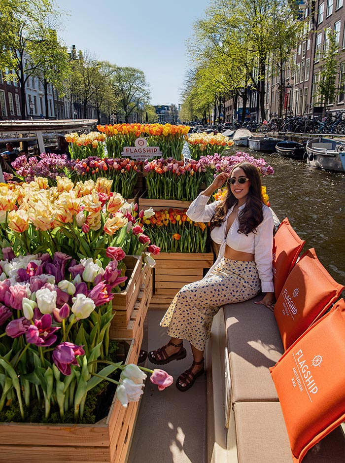 Woman enjoying a tulip boat cruise with Flagship Amsterdam, seated among wooden crates overflowing with colorful tulips in shades of pink, yellow, orange, and white. She wears sunglasses, a white top, and a floral skirt, smiling as the boat cruises along a scenic canal lined with historic buildings and springtime trees. The orange seat cushions add a cozy, branded touch.