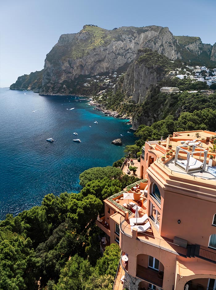 Breathtaking view from Hotel Punta Tragara in Capri, Italy, with its terracotta building perched on a cliffside overlooking lush greenery, turquoise waters, and dramatic coastal cliffs dotted with white villas and boats.