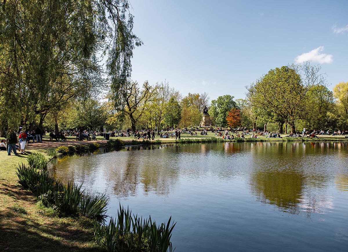 Sunny day at Vondelpark in Amsterdam with people relaxing, walking, and picnicking on the grassy lawns beside a calm pond. Trees in various shades of green and orange frame the scene, and a statue is visible in the distance. The water reflects the foliage and blue sky, creating a peaceful urban oasis.







