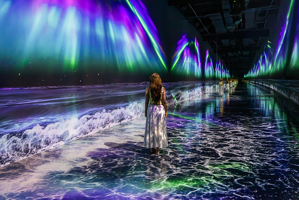 A woman in a long white skirt walks through an immersive art exhibition featuring projections of crashing ocean waves and glowing purple green and blue northern lights. The colorful projections stretch along the walls and reflect across the floor of a long dark gallery.