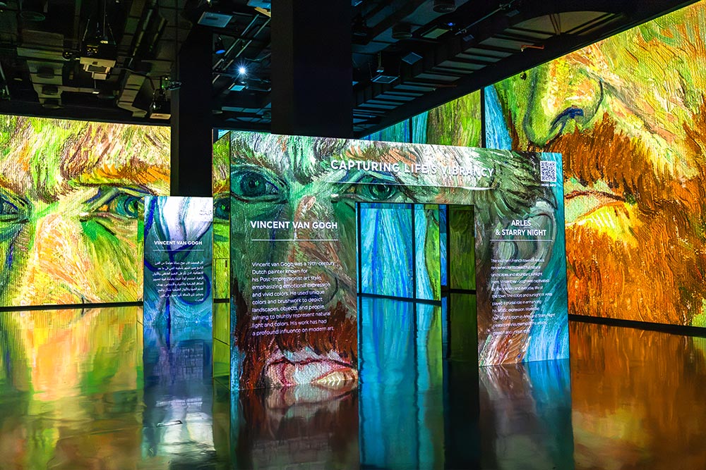 An immersive Vincent van Gogh exhibition featuring enormous colorful projections of his self portrait across the gallery walls. An illuminated entrance titled "CAPTURING LIFE'S VIBRANCY" displays information about Van Gogh and Arles with the vibrant green blue and orange artwork reflected across the glossy floor.
