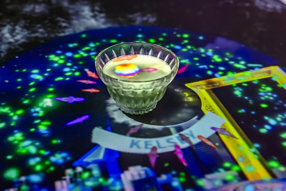 A small glass bowl filled with a pale green cocktail sits on a table covered in colorful projected lights and animated fish. A white banner reading KELSEY is surrounded by glowing blue and green patterns.