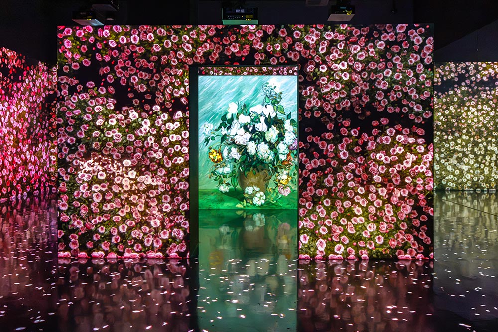 An immersive art exhibition with walls covered in projections of pink and red flowers surrounding a central display of a painted bouquet of white flowers in a vase. The colorful floral projections reflect across the glossy floor of the dark gallery.