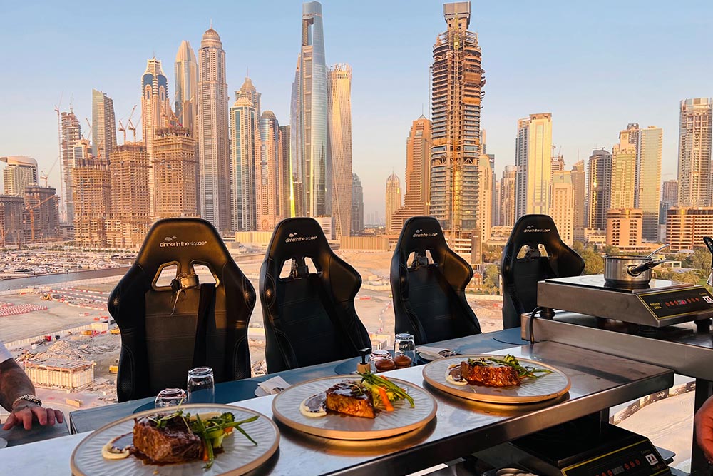 Three plated meals sit on the dining counter of a suspended Dinner in the Sky platform overlooking the Dubai Marina skyline. Black dining seats face towering skyscrapers illuminated by the warm golden light of the setting sun.