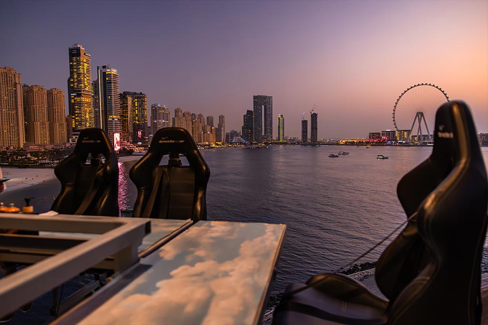 Empty seats and dining tables on a suspended Dinner in the Sky platform overlooking Dubai Marina at sunset. The illuminated skyscrapers of JBR and Ain Dubai observation wheel rise above the waterfront beneath a pink and purple evening sky.