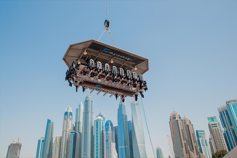 A group of diners sits in a suspended Dinner in the Sky platform high above the skyscrapers of Dubai Marina. The open air dining platform hangs from a crane against a clear blue sky with the Dubai skyline in the background.
