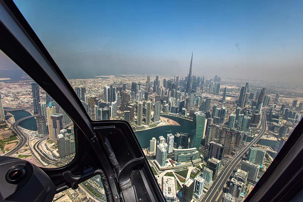Aerial view of Downtown Dubai from inside a helicopter. The Burj Khalifa towers above the surrounding skyscrapers with winding roads and waterways visible below during a scenic helicopter tour.
