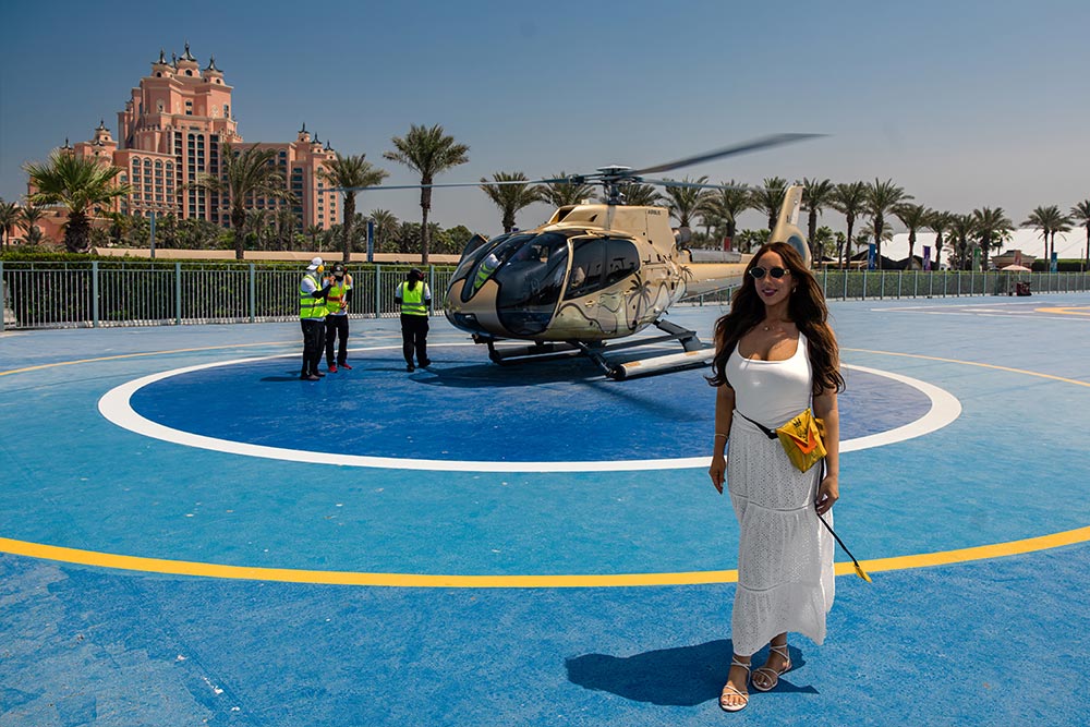A woman in a white summer dress and sunglasses poses in front of a helicopter on a blue helipad at Atlantis The Palm in Dubai. The pink hotel and rows of palm trees stand in the background beneath a clear blue sky.