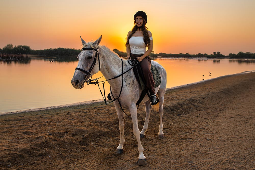 A woman wearing a black riding helmet sits on a white horse beside a calm lake at sunset. The golden orange sky reflects across the water behind her as she enjoys a sunset horseback ride along the sandy shore.