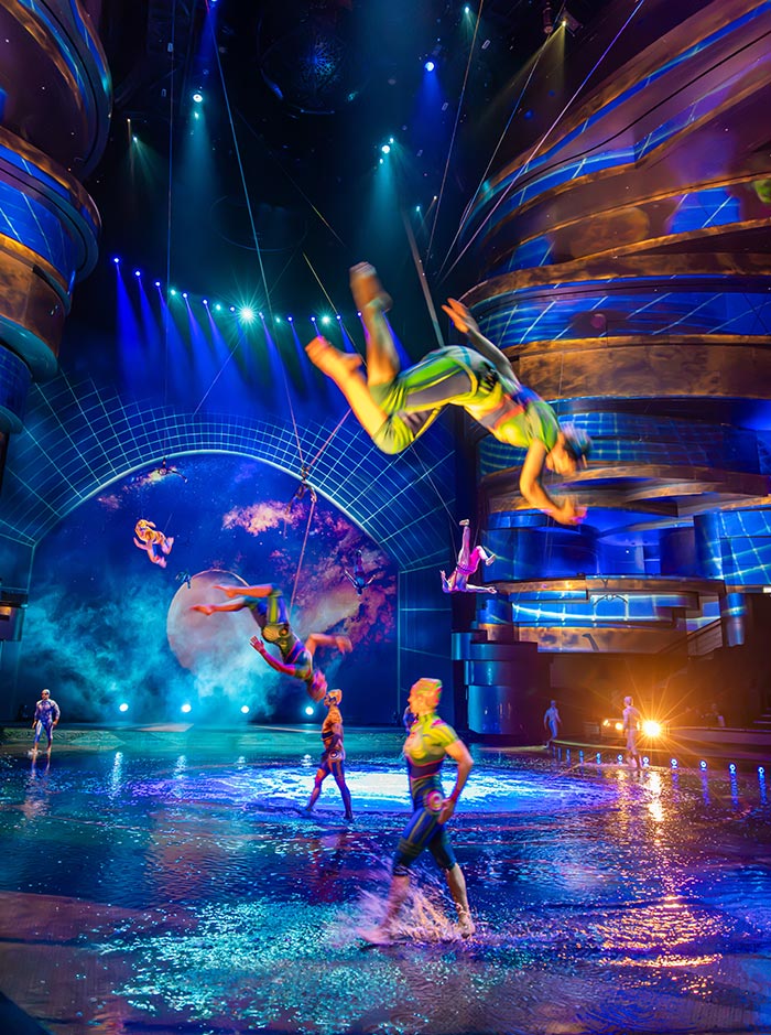 Acrobats in colorful costumes perform aerial flips and stunts above a water covered stage during an indoor live show. Blue and purple spotlights illuminate the performers as others splash through the shallow water beneath towering futuristic structures.