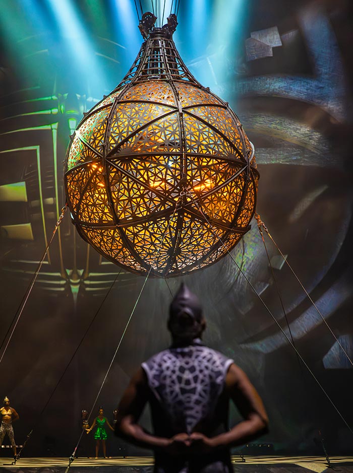 A large spherical metal cage with intricate geometric patterns hangs above a stage illuminated by warm golden light and blue spotlights. A performer stands beneath the suspended structure while other performers wait in the background during a theatrical acrobatic show.