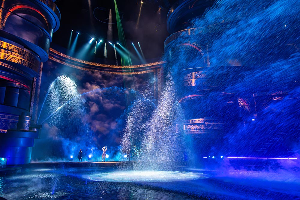 Performers stand onstage during a spectacular indoor water show as enormous fountains shoot arcs of water across the stage. Bright blue spotlights illuminate the cascading water and towering circular structures decorated with intricate golden patterns.