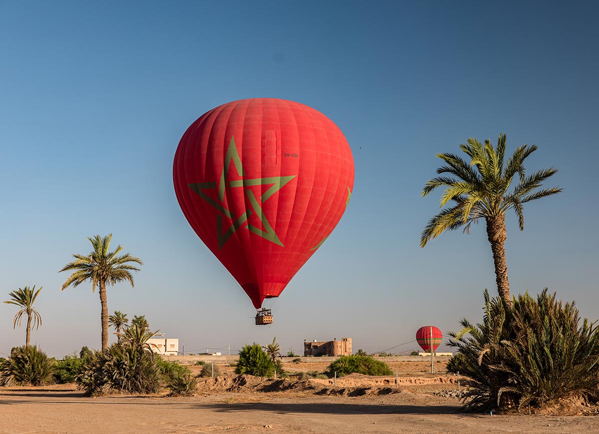A red hot air balloon with a green Moroccan star floats above a palm-lined desert landscape, with a second balloon in the distance beneath a clear blue morning sky.