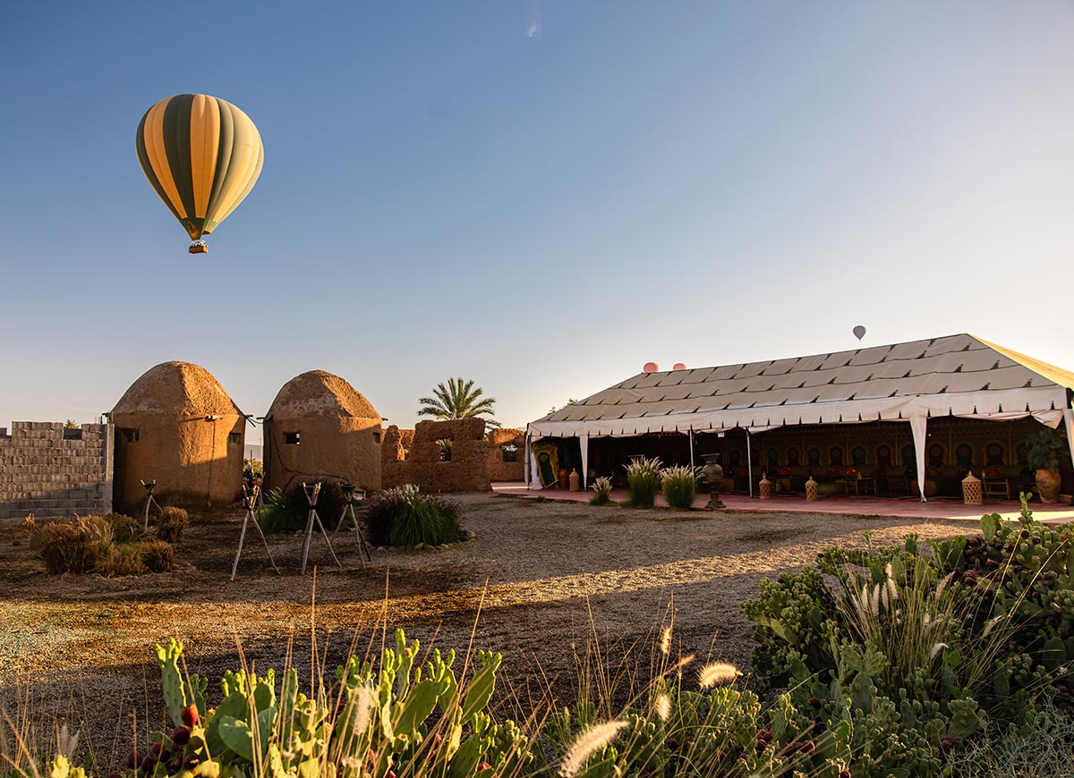 A hot air balloon floats above a traditional desert camp with earthen huts and a large Moroccan tent, surrounded by cacti and palm trees in the warm morning light.
