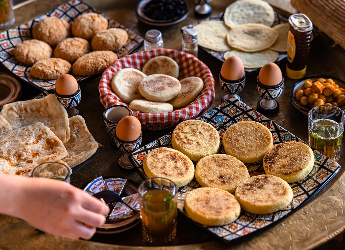 A traditional Moroccan breakfast spread with freshly baked breads, msemen, baghrir, boiled eggs, olives, chickpeas, honey, mint tea, and assorted preserves served on colorful patterned platters.