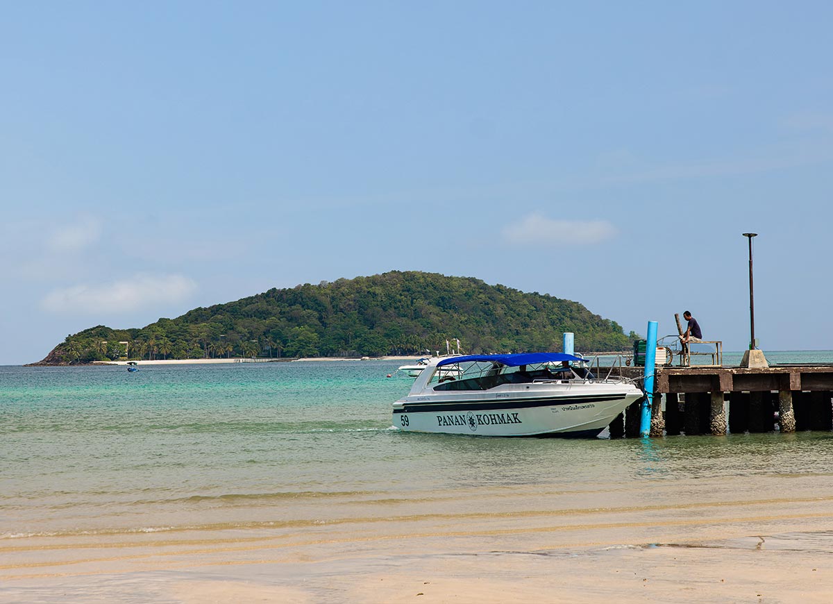 Speedboat docked at a small pier beside a sandy beach with shallow clear water in the foreground. A person stands on the pier while a green island rises in the background under a bright blue sky.