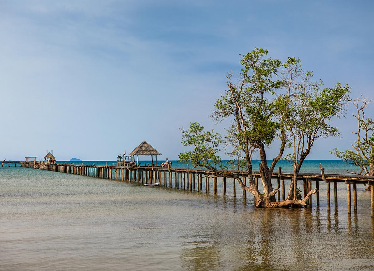 Long wooden pier stretching across shallow calm water with a small thatched pavilion at the end. A tree grows beside the pier while the ocean and distant islands sit under a soft blue sky.