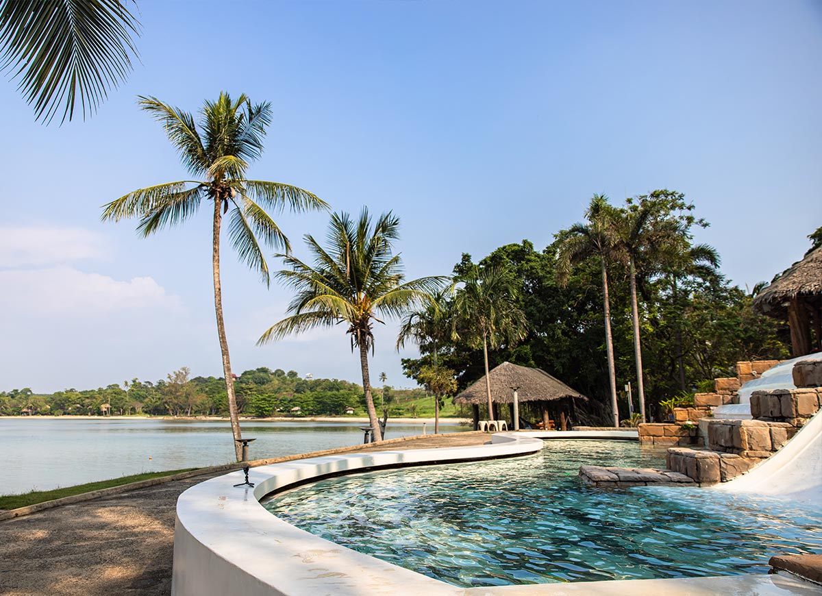 Curved resort pool with clear blue water beside a calm lagoon lined with palm trees and lush greenery. A small thatched hut and stone water feature create a relaxing tropical setting under a clear sky.