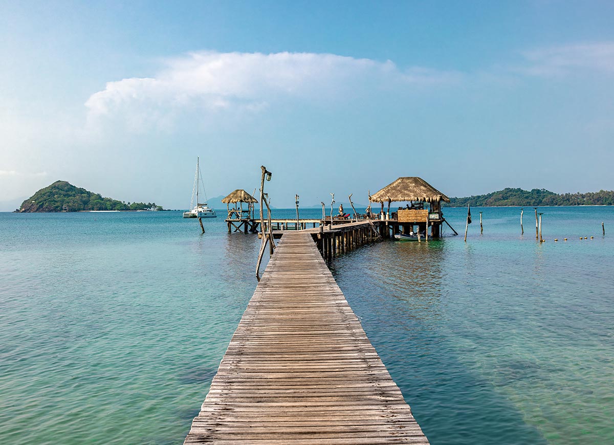 Long wooden pier stretching over clear turquoise water toward a small thatched roof bar on stilts. A sailboat floats nearby and green islands sit on the horizon under a bright blue sky.