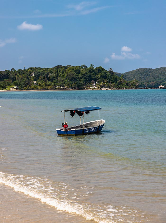 Small blue boat with a canopy floats in shallow clear water near the shore, with the words "KOH KHAM" painted on its side. Gentle waves lap at the sandy beach in the foreground while a lush green island stretches across the horizon under a bright blue sky.