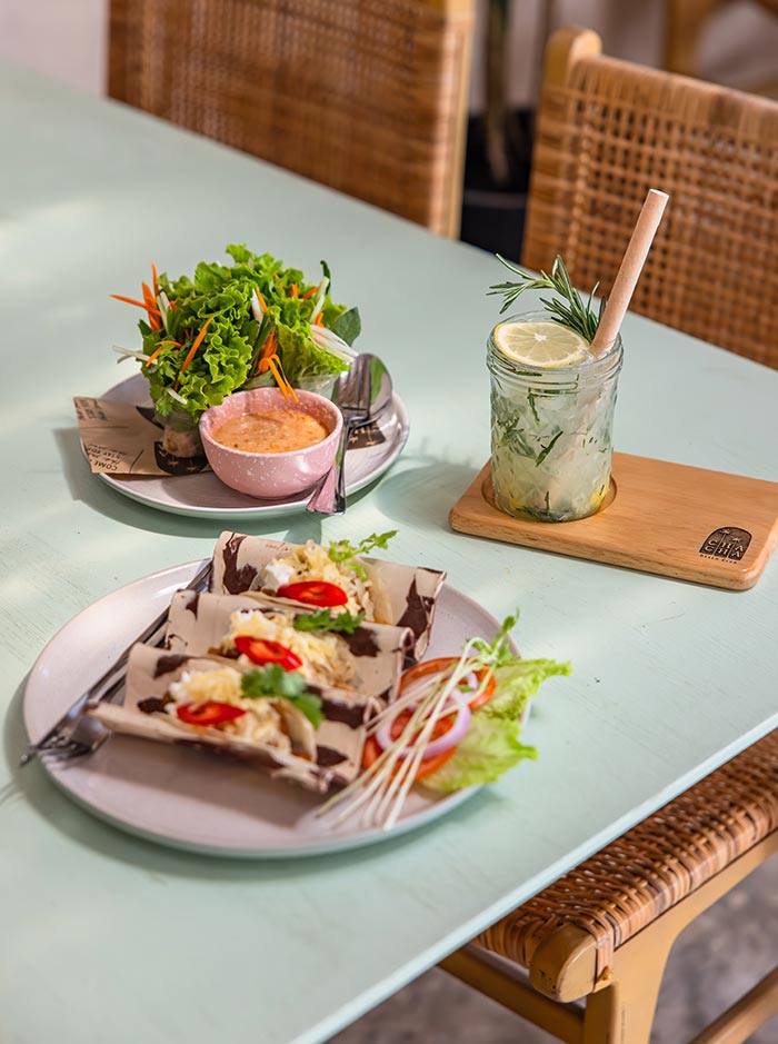 Plate of tacos topped with shredded cabbage and red chili slices served with a side of fresh greens and dipping sauce on a pastel table. A glass of iced herbal drink with lemon and rosemary sits nearby on a wooden coaster beside woven chairs.