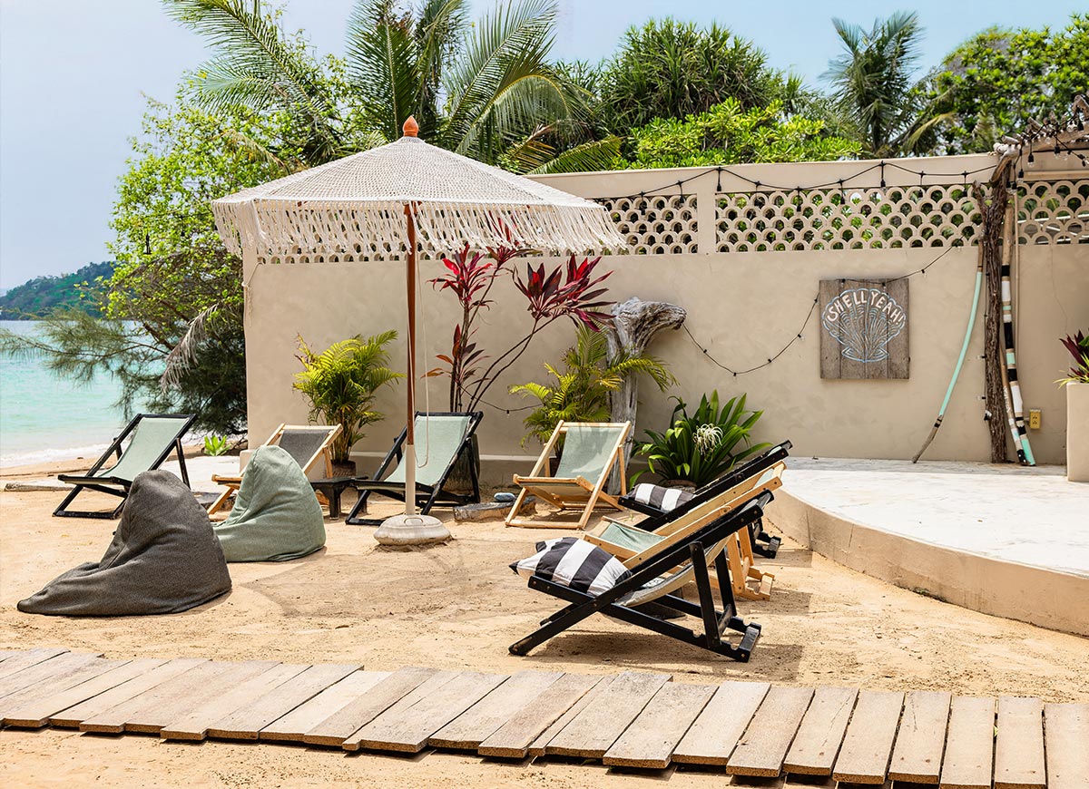 Sandy beach seating area with wooden lounge chairs and bean bags arranged around a fringed umbrella near the ocean. A cream wall with string lights and a sign reading "SHELL YEAH" sits behind tropical plants and palms.