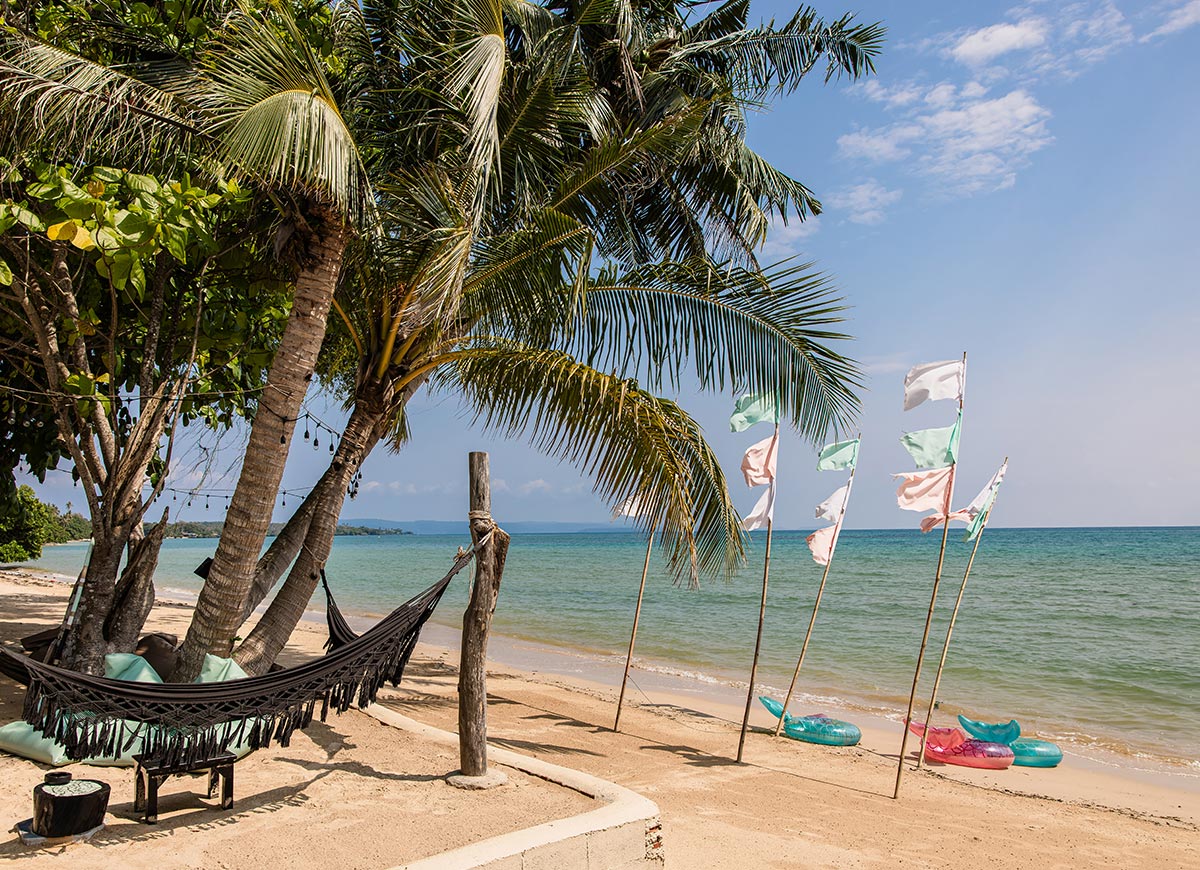 Sandy tropical beach with a hammock strung between palm trees facing calm turquoise water. Colorful flags line the shore while inflatable rings rest on the sand under a bright blue sky.