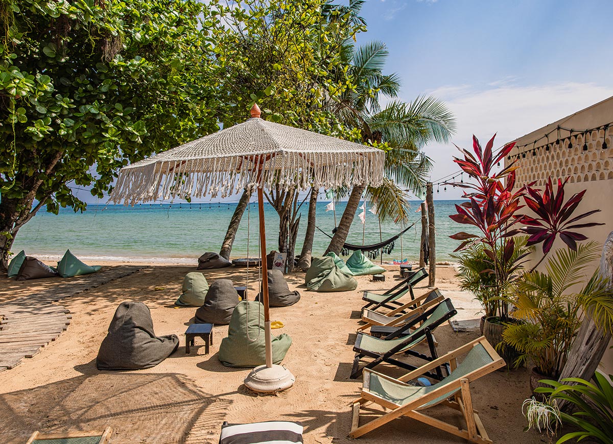 Sandy beach lounge with wooden chairs and bean bags arranged under a fringed umbrella and palm trees beside the ocean. Calm turquoise water stretches behind the seating area with a hammock and string lights adding to the relaxed tropical setting.