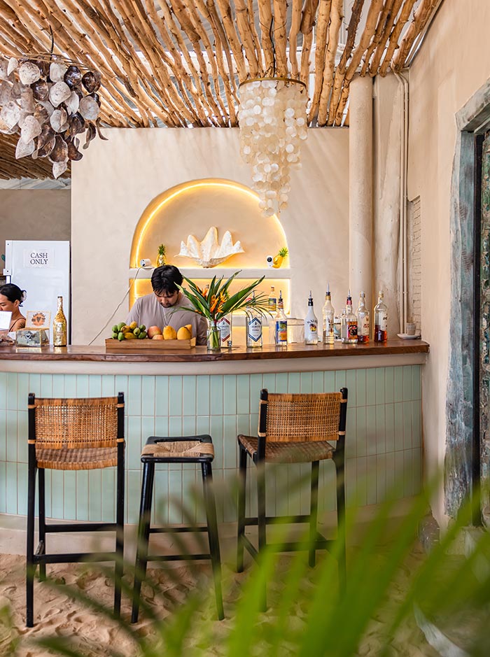 Beach bar with a wooden counter and woven chairs set on sand beneath a ceiling of bamboo sticks. A bartender prepares drinks beside bottles and fresh fruit while a warm backlit shelf with seashell decor glows against a neutral wall.