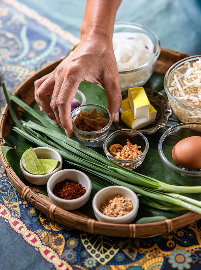 Hand reaching for small bowls of ingredients arranged on a tray lined with a banana leaf, including chopped herbs, dried shrimp, chili flakes, and crushed peanuts. Fresh green onions, tofu, egg, lime wedges, and noodles are laid out for preparing a stir fried noodle dish.