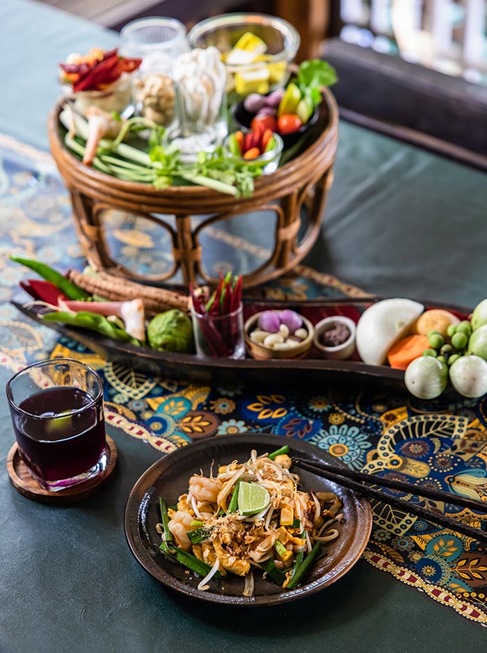 Plate of stir fried noodles with shrimp, bean sprouts, and lime served with chopsticks on a patterned table. Fresh herbs, vegetables, and spices are arranged in trays nearby with a glass of dark drink completing the meal setup.