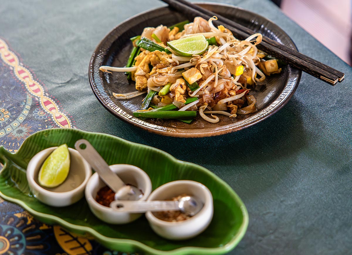 Plate of stir fried noodles with tofu, bean sprouts, green onions, and a lime wedge served with chopsticks. Small bowls of condiments and lime sit on a leaf shaped dish beside the meal on a patterned table.