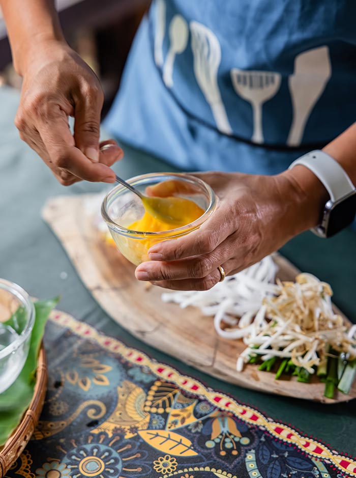 Hands whisking beaten egg in a small glass bowl beside fresh noodles, bean sprouts, and chopped greens on a wooden board. A person wearing a blue apron prepares ingredients for a stir fried noodle dish on a patterned table.