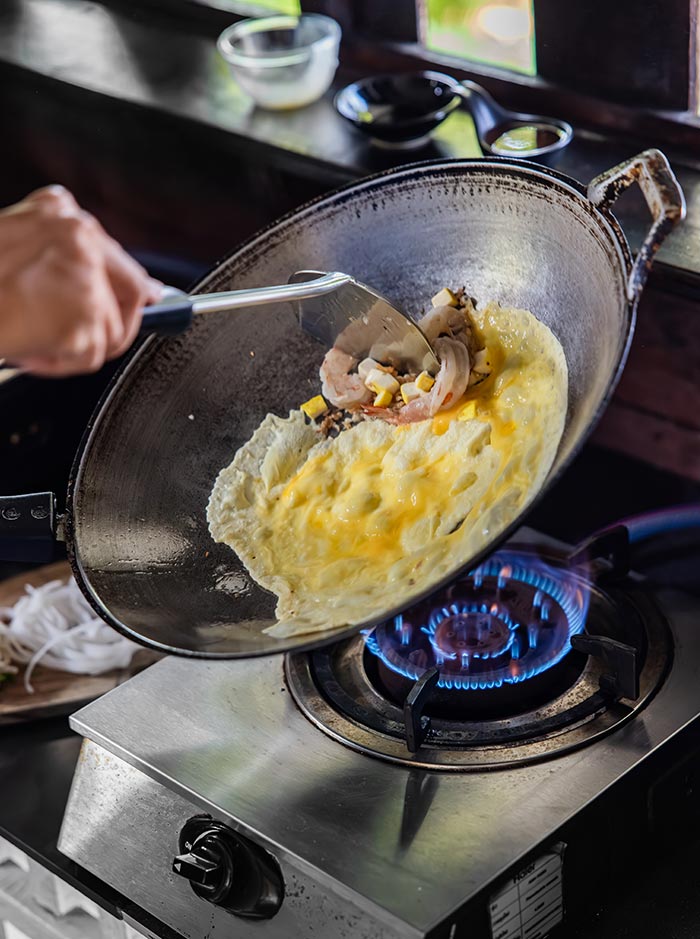 Egg cooking in a hot wok over a blue gas flame as a hand uses a spatula to fold it together with shrimp and diced ingredients. The scene captures the start of stir frying noodles with fresh ingredients visible nearby.
