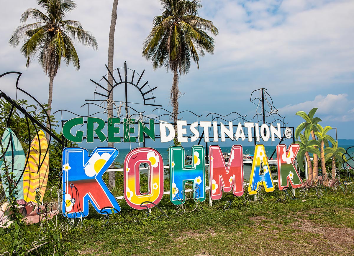 Colorful outdoor sign reading "GREEN DESTINATION KOH MAK" decorated with bright patterns, surfboards, and tropical illustrations. Palm trees and ocean views appear behind the sign under a partly cloudy sky.