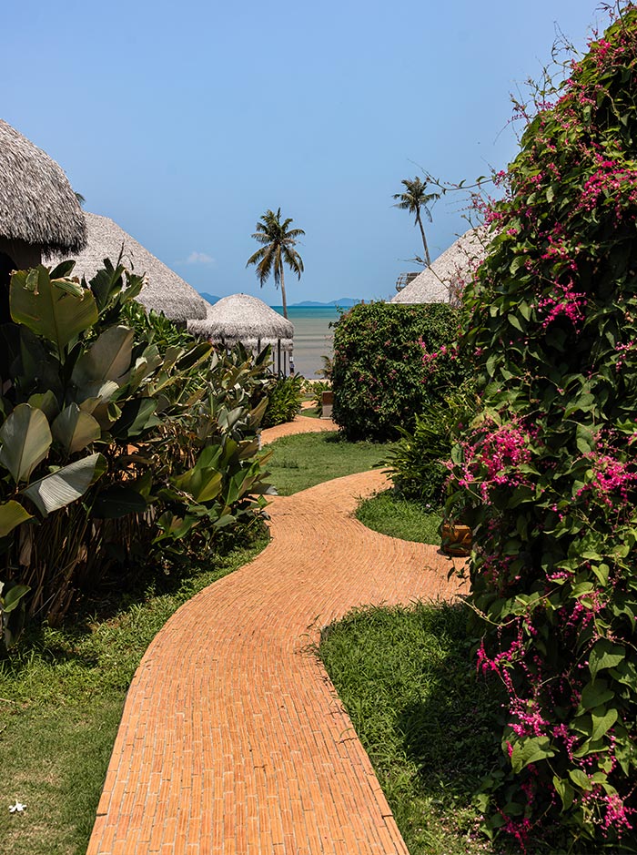 A curved brick pathway leads through lush tropical greenery and flowering bushes toward thatched roof huts and the ocean beyond. Palm trees and vibrant plants frame the scene, guiding the view to the calm blue water in the distance.