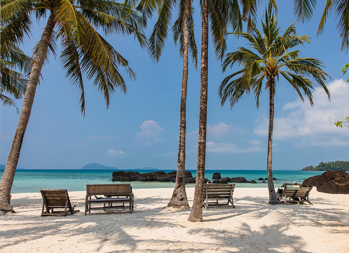 Wooden lounge chairs sit beneath tall palm trees on a white sandy beach, facing calm turquoise water dotted with dark rocks. A distant island and clear blue sky complete the peaceful tropical scene.