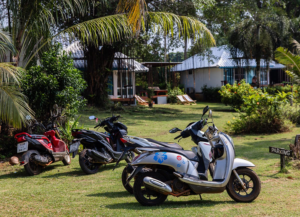 Three motorbikes parked on grass beneath palm trees in a tropical garden setting. Small bungalow style cottages and lounge chairs sit in the background near a sign that reads "PARKING".