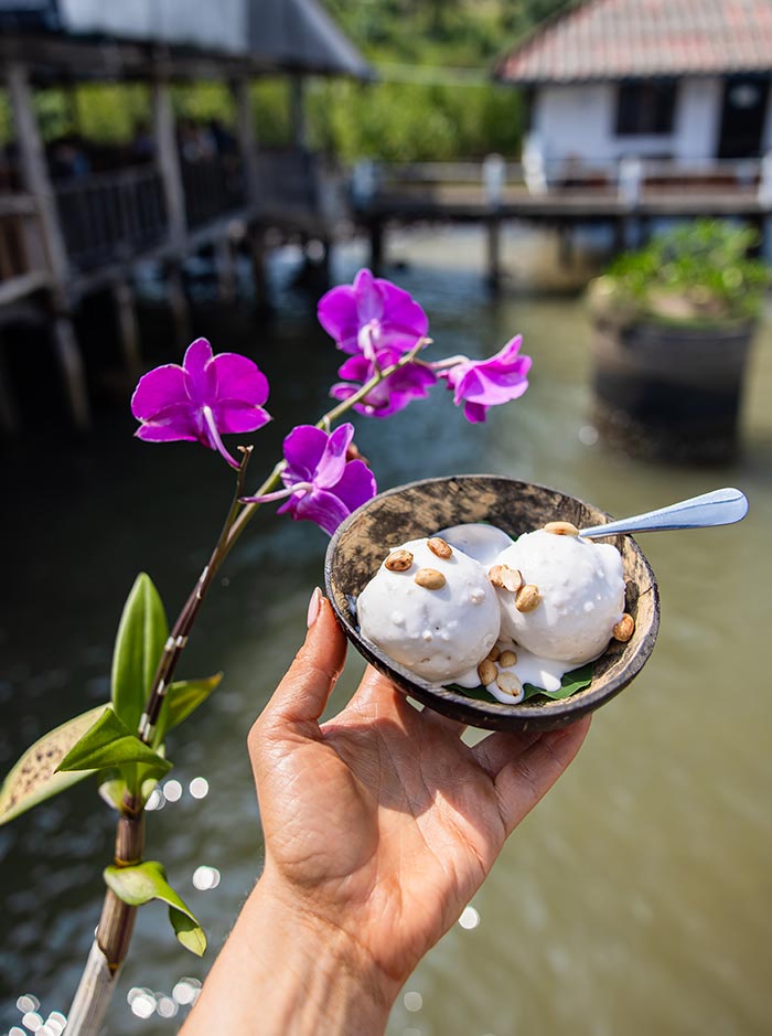Hand holding a small bowl of coconut dessert topped with peanuts beside a purple orchid flower over calm water. Wooden buildings on stilts blur in the background, creating a relaxed tropical setting.