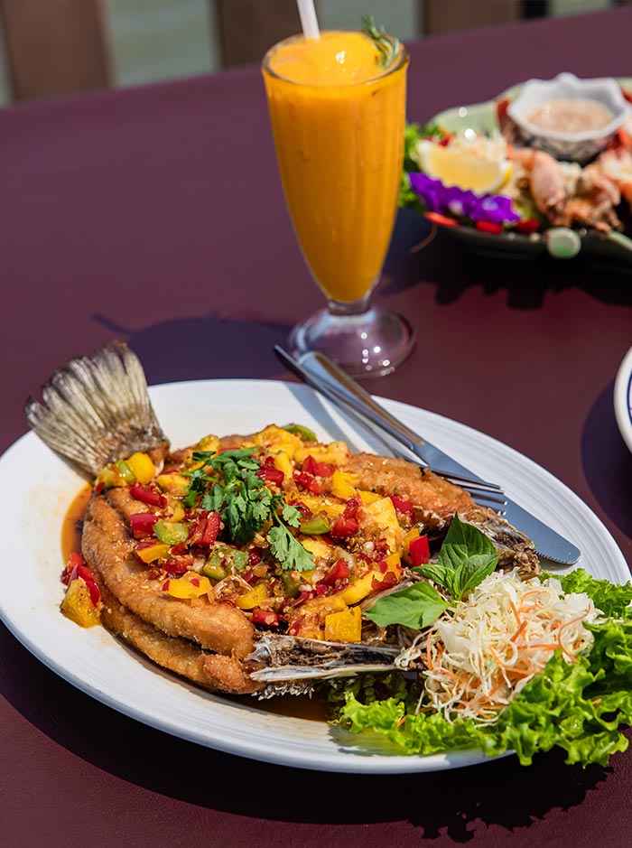 Plate of fried whole fish topped with colorful chopped peppers, herbs, and sauce served with fresh greens on the side. A tall glass of bright orange smoothie sits behind the dish on a table.