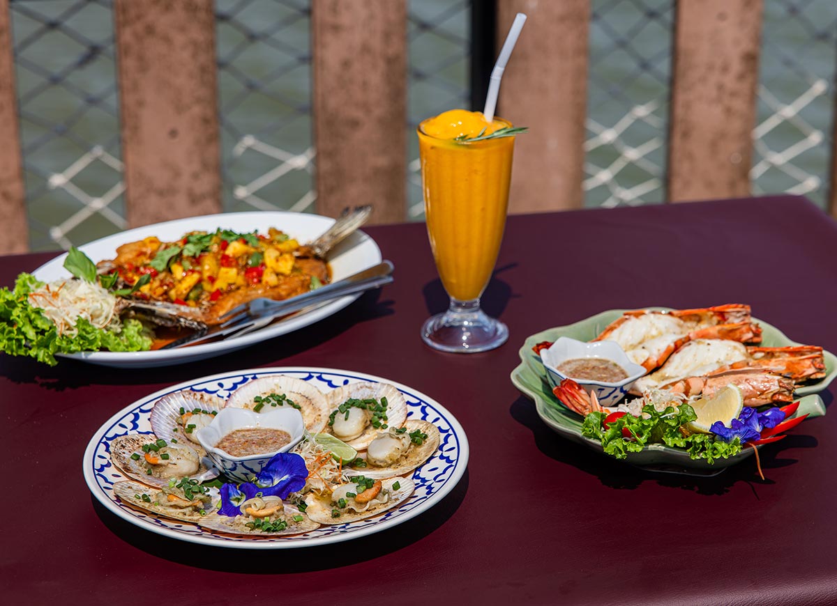 Three plates of seafood dishes including scallops in shells, grilled prawns, and a whole fried fish topped with colorful vegetables arranged on a table. A tall orange smoothie sits in the center with water visible through a railing in the background.