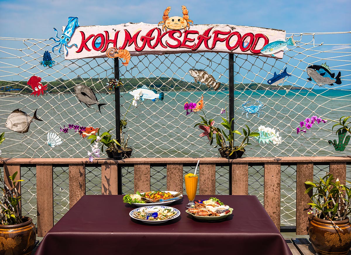 Table set with seafood dishes and a bright orange smoothie in front of a seaside railing decorated with colorful fish illustrations. A sign above reads "KOH MAK SEAFOOD" with the ocean stretching out in the background.