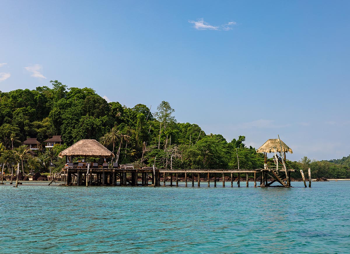 A wooden pier stretches over clear turquoise water to two thatched roof huts, backed by a lush green tropical hillside. The calm sea and bright blue sky create a peaceful island setting with rustic structures perched above the water.