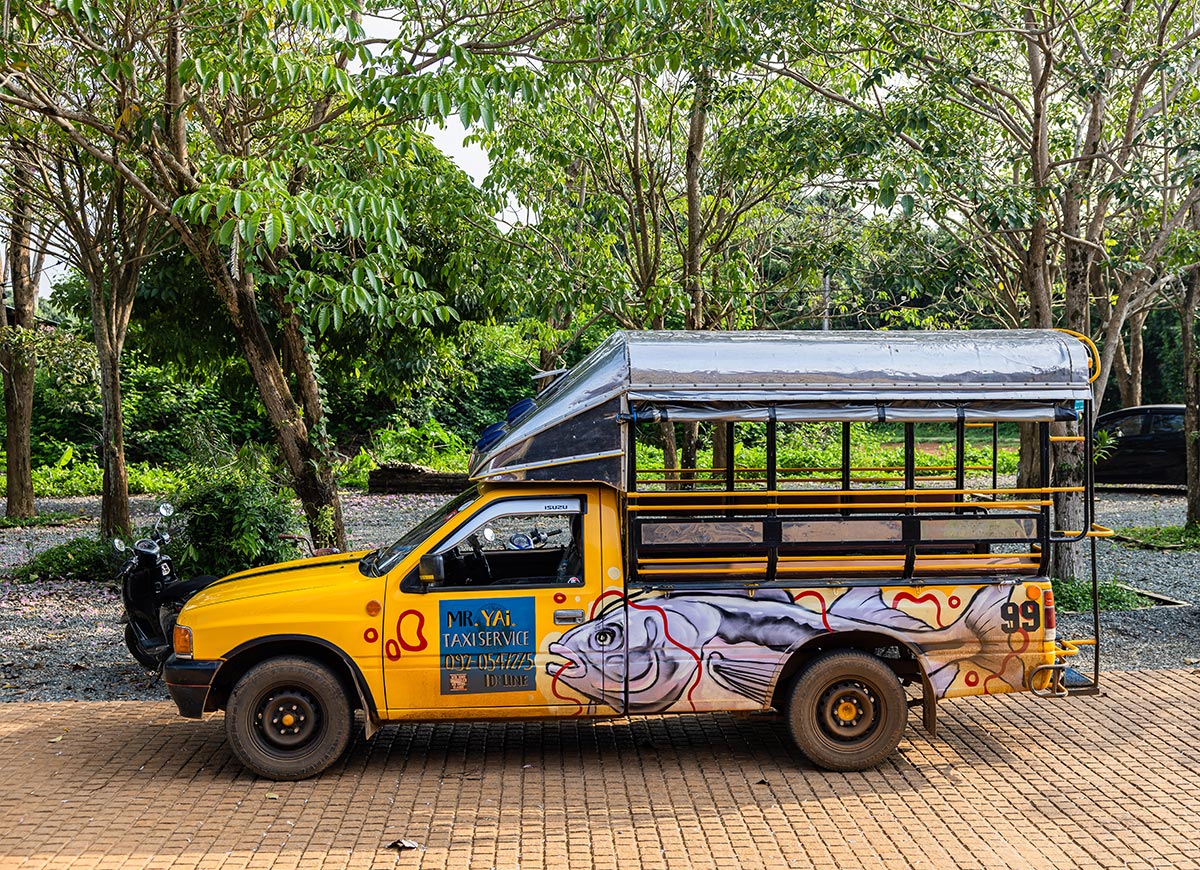 Yellow open air taxi truck with bench seating parked on a paved area surrounded by trees. A painted fish decorates the side along with text reading "MR. YAI TAXI SERVICE 087 054 4475 ID LINE" on the door.