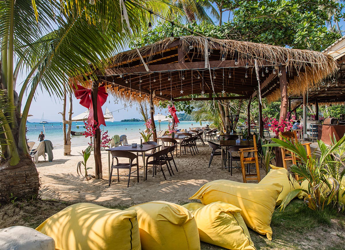 Open air beachfront restaurant with wooden tables and chairs set on sand beneath a thatched roof decorated with string lights and red bows. Palm trees frame the space while the ocean and anchored boats sit just beyond the seating area.