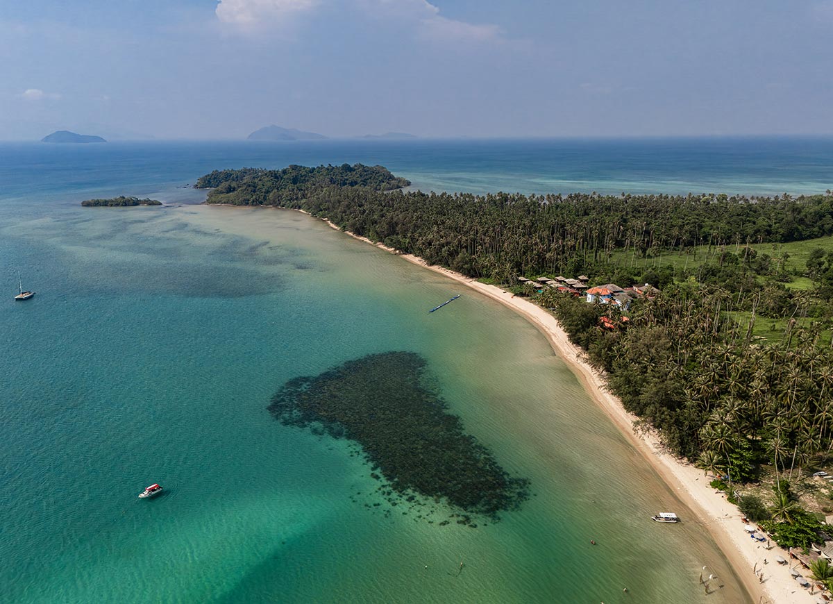 Aerial view of a long curved sandy beach bordered by dense palm trees and clear turquoise water. Dark reef patches appear beneath the surface while small boats float offshore and distant islands fade into the horizon.