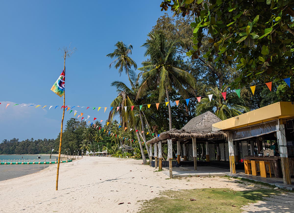 A beachfront bar with a thatched roof sits along a sandy tropical shore lined with palm trees, decorated with colorful triangular flags strung overhead. A flag on a tall pole waves beside the water while calm sea and a tree-covered coastline stretch into the distance under a clear blue sky.