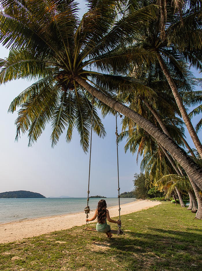 Person sitting on a rope swing hanging from a tall palm tree beside a quiet sandy beach. Calm turquoise water and distant islands create a peaceful tropical backdrop.