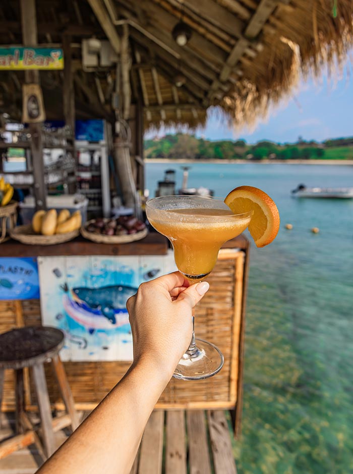 Hand holding a cocktail in a stemmed glass garnished with an orange slice in front of a thatched beach bar over clear water. The bar with stools and fruit display sits in the background while a boat floats on the calm sea.